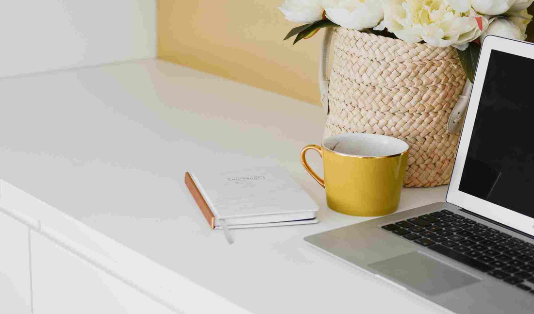 bureau avec un mug jaune un pot de fleur en osier avec des fleurs blanches un carnet et un ordinateur portable 